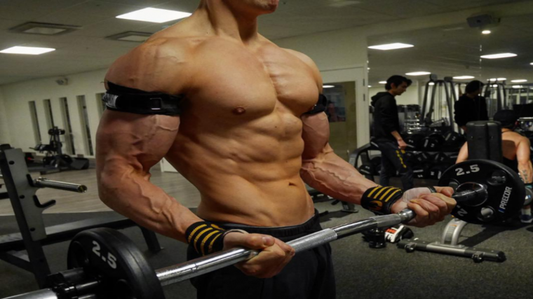 A person performs a barbell curl while restricting their blood flow with cuffs.