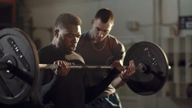 A person performs a barbell curl with their coach nearby.