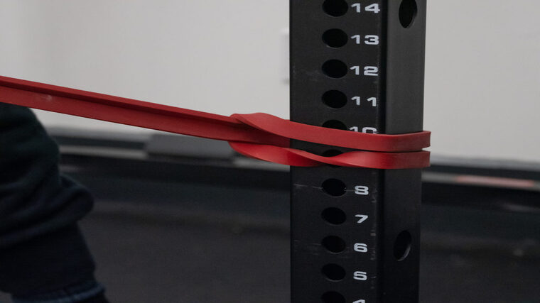A close up of a red resistance band looped securely around a squat rack leg.