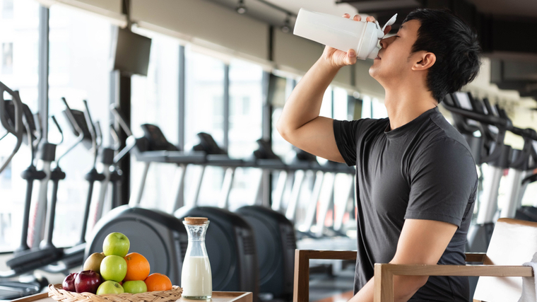 A person drinks a protein shake next to a plate full of fruit in the gym.