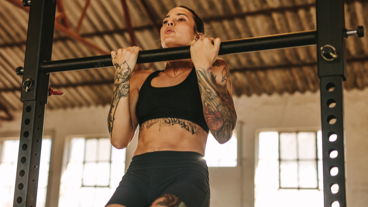 A person with tattoos on both arms wears a sports bra while performing a chin-up.