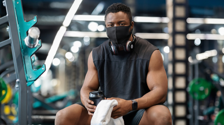 A person sits in the gym while wearing a mask with headphones around their neck.
