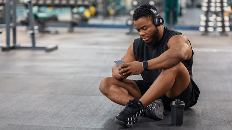 A person sits on the floor in the gym while listening to headphones and looking at their phone.