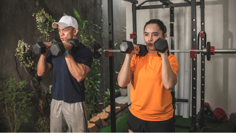 Two people wearing t-shirts and workout shorts hold dumbbells ready to press overhead.