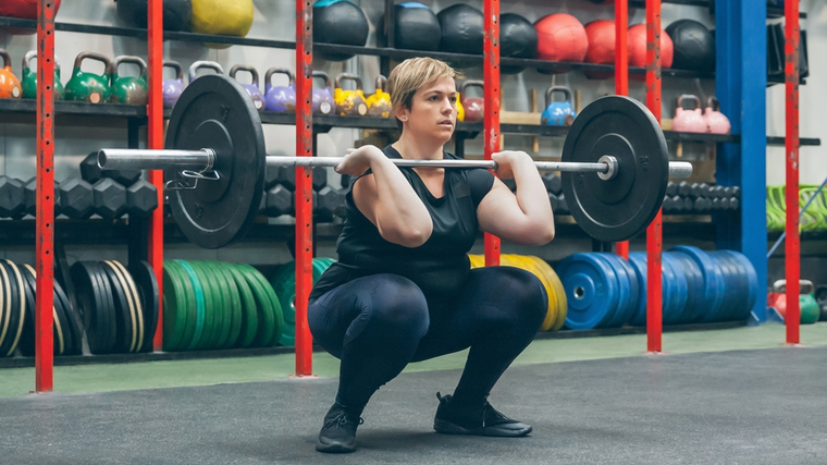 A person wearing a black t-shirt and workout pants performs a front squat with a loaded barbell.