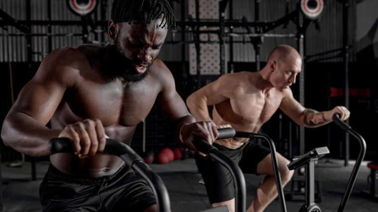 Two shirtless people work on air bikes next to each other.