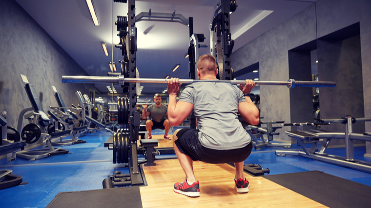 A person is featured from behind, wearing a grey t-shirt performs a squat with an empty barbell.