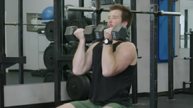 An athlete wearing a black tank top holds two dumbbells facing toward him at chin level while sitting on a bench.