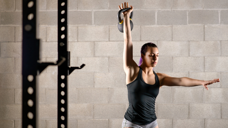 A person presses one kettlebell overhead with their other arm spread out for balance.