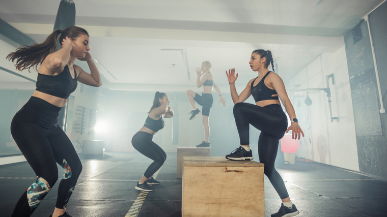 Four people perform either jump squats or plyo box step-ups in a fitness studio.