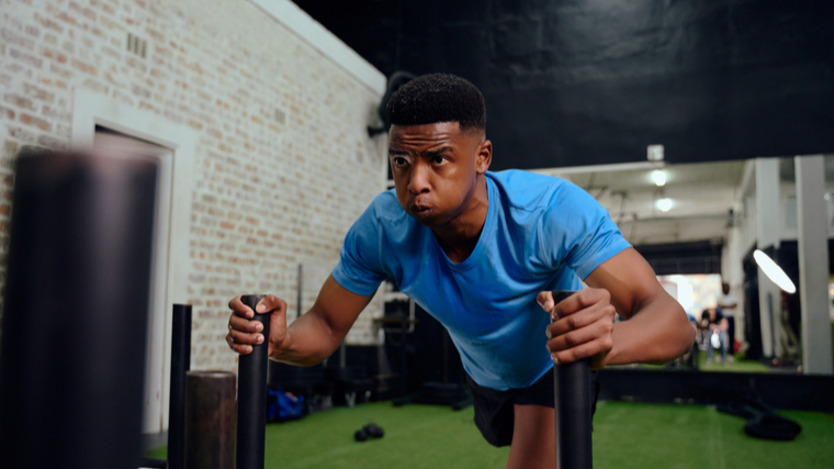 A person wears a blue t-shirt and exhales heavily while pushing a sled in the gym.