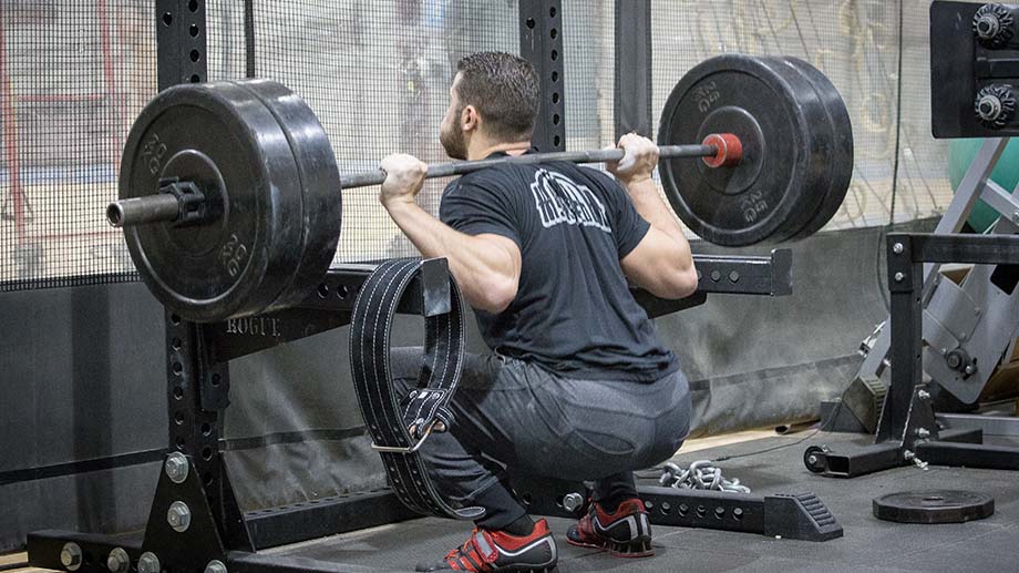 Man in a black t-shirt and black pants squatting deep with a loaded barbell across his back.