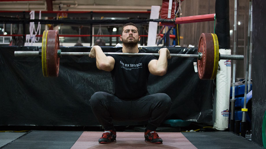 Man in black t-shirt and pants sitting in a deep squat position holding a loaded barbell in a front-rack position.