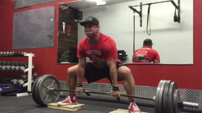 A person wearing a red Westside Barbell shirt, lifting belt, and baseball cap prepares to deficit sumo deadlift.
