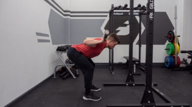 A person wearing a red t-shirt and black pants performs a good morning with a barbell.