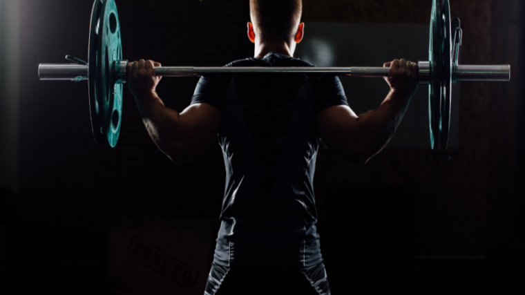 A person facing away from the camera prepares to perform a low bar back squat.