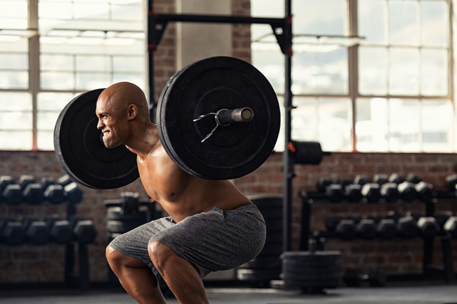 A shirtless person scrunches their face with effort as they perform a barbell back squat in the gym.