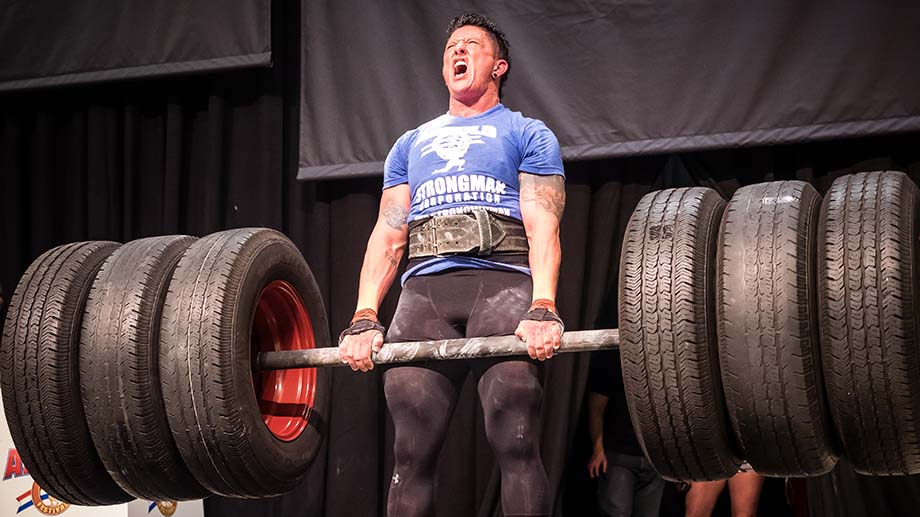 Strongman competitor, wearing black leggings, a weightlifting belt, and a blue t-shirt, deadlifting hummer tired attached to a barbell