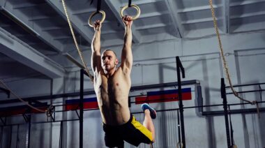 Man in yellow shorts and no t-shirt doing muscle-ups on a pair of rings.
