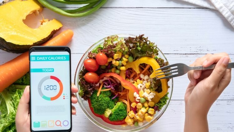 A calorie tracker app on a mobile phone on one hand, and a fork on another for the food on the table.