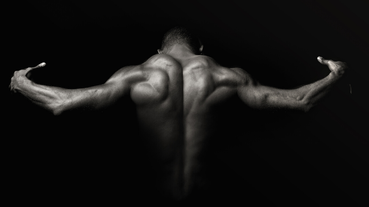 An athlete in a dark room flexing back and shoulder muscles with light accenting muscle form and build.
