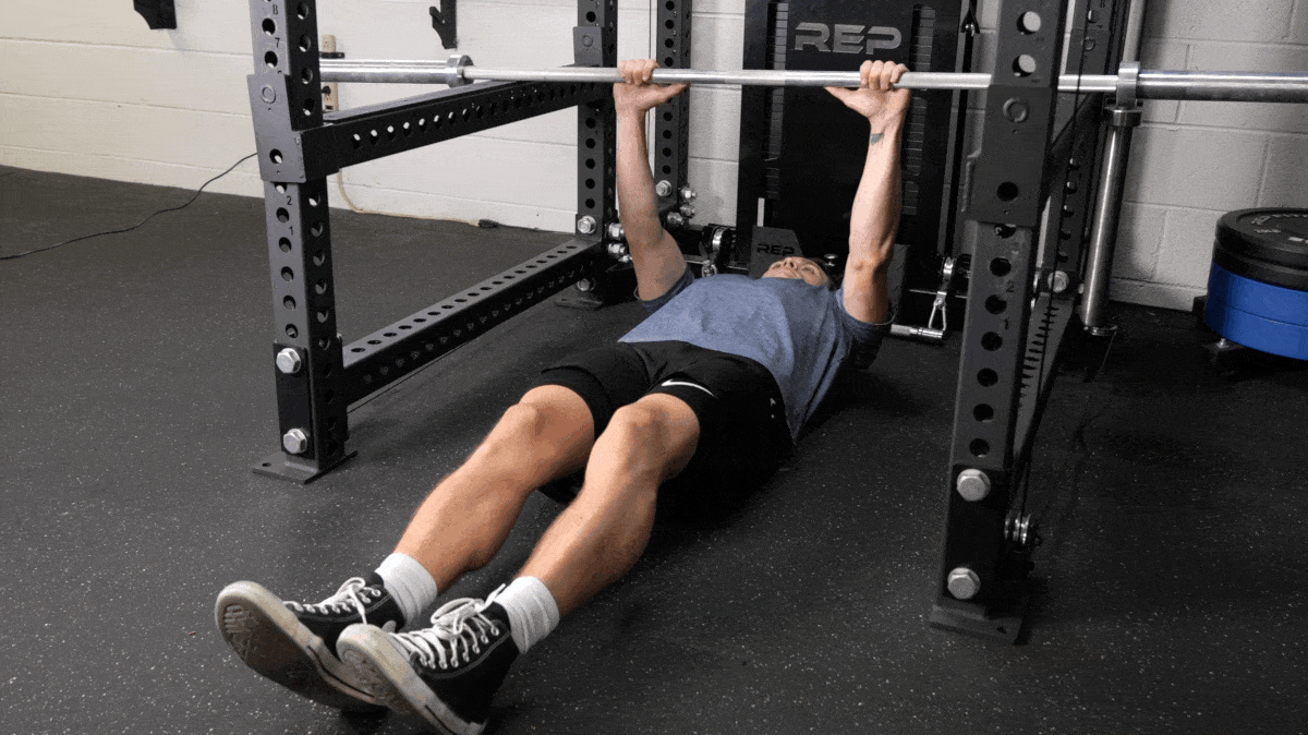 A person wearing black shorts and blue t-shirt performing an inverted row with a barbell set in a power rack.