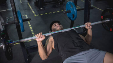 A person wears a black tank top and holds a bench press at the bottom of the lift.