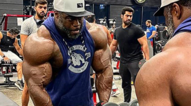 Brandon Curry wears a baseball cap and muscle tank while lifting weights.