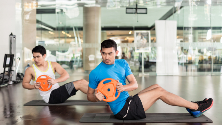 Two people perform Russian twists in the gym.