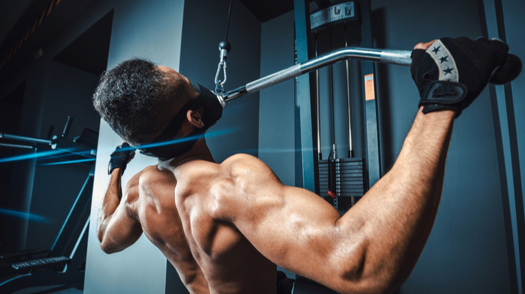 A shirtless person wears a mask and workout gloves while performing a lat pulldown.