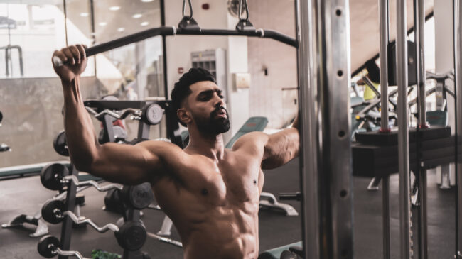 A person performs a lat pulldown shirtless in a gym.