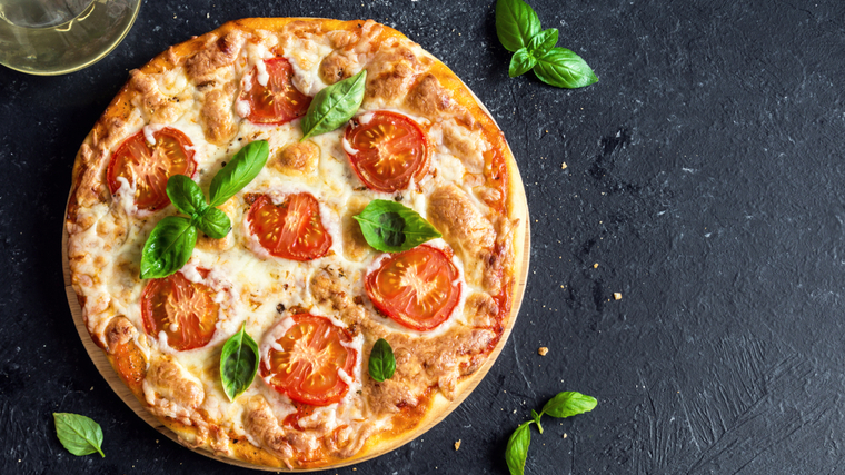 A pizza with fresh basil and tomato slices sits on a black tabletop.