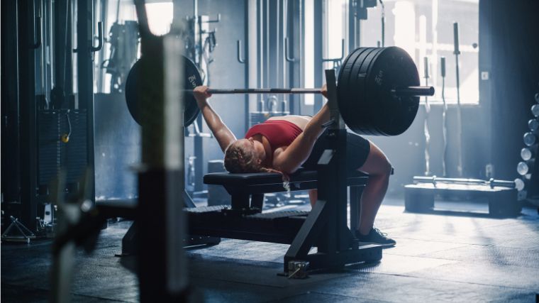 person in a red tank top, arching their back on a training bench while holding a barbell loaded with bumper plates, about to bench press the weight.
