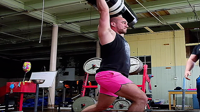 A person wearing pink shorts locks out a log bar overhead using a jerk technique.