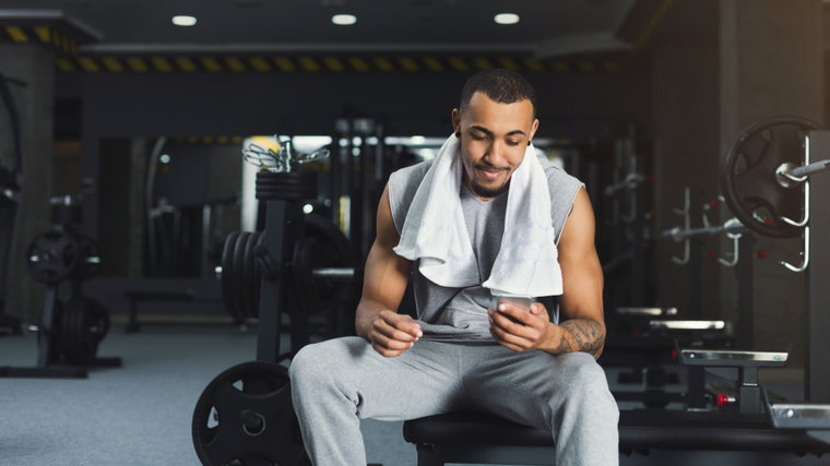 A person with a towel draped over their shoulder sits on a bench in the gym and looks at their phone.