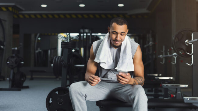A person with a towel draped over their shoulder sits on a bench in the gym and looks at their phone.