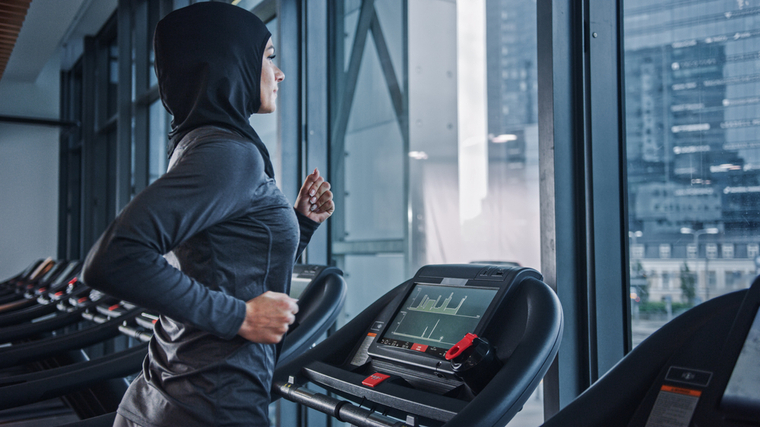 A person wearing a hijab and a grey shirt runs on a treadmill.