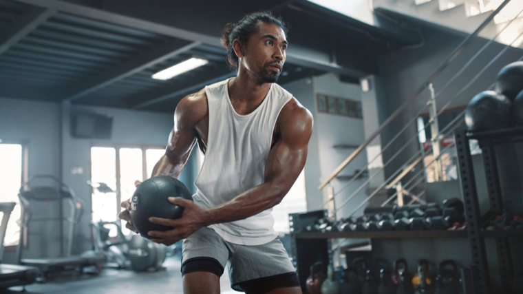 A person with their hair pulled back wearing a white tank top holds a medicine ball at their side in an athletic stance.
