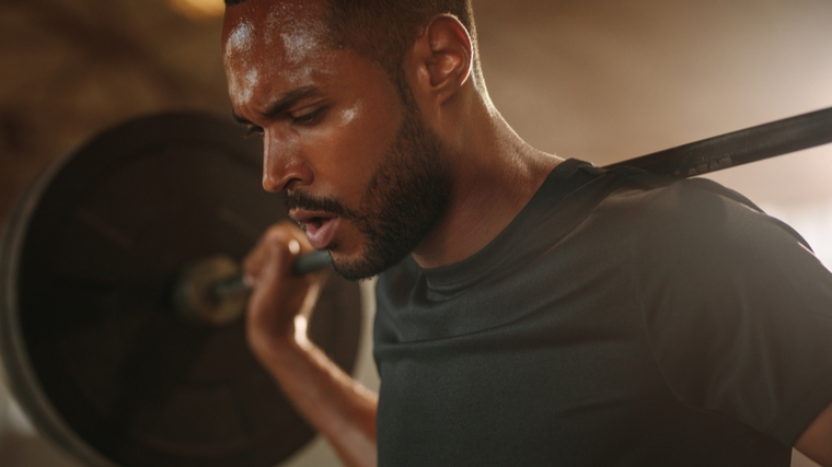 A person with facial hair sweats and looks down with a loaded barbell on their back.