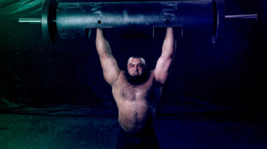 A shirtless, bearded person performs a log press.