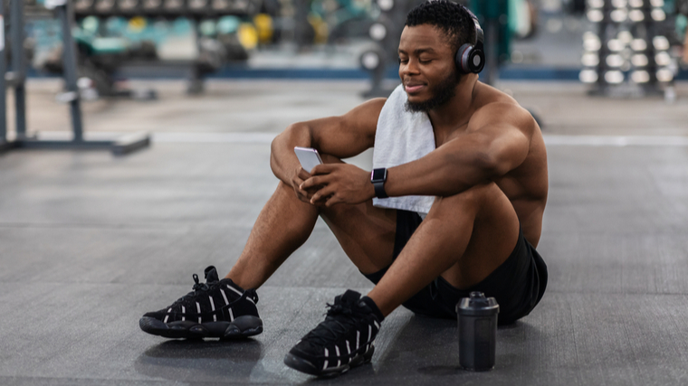 A shirtless person with a towel on their shoulder wears headphones while sitting on the gym floor, looking at their smartphone.