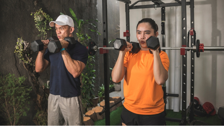 Two people in T-shirts and workout shorts have dumbbells ready to push overhead.