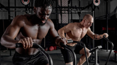 Two shirtless people work on air bikes next to each other.