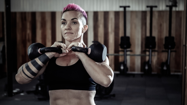A person with short pink hair and forearm tattoos wears a sports bra while holding two kettlebells in front rack position.