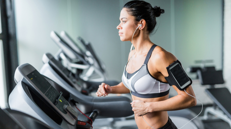 A person wearing a grey sports bra looks ahead while wearing headphones and a smartphone in their armband.