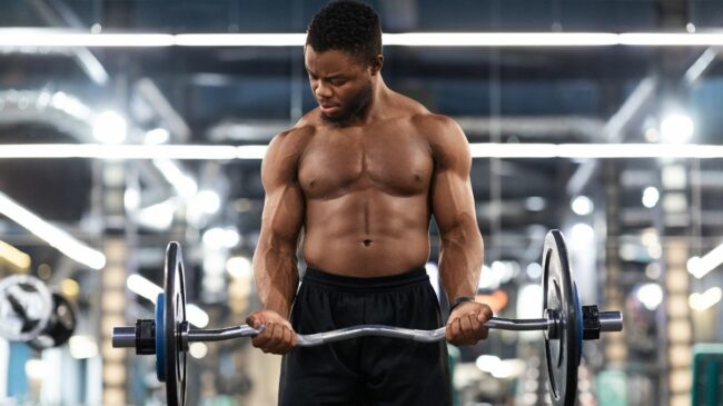 A dark-skinned person wearing shorts and no shirt curls a loaded barbell in a gym