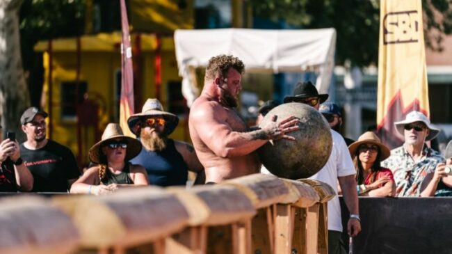 Strongman Tom Stoltman holds an Atlas Stone, shirtless, on top of a wooden beam in front of fans at the 2021 World's Strongest Man contest