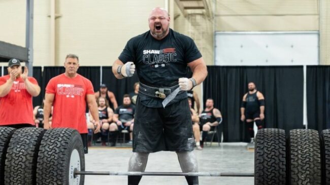 Strongman Brian Shaw standing in front of a barbell loaded with Hummer tires screaming with clenched fists