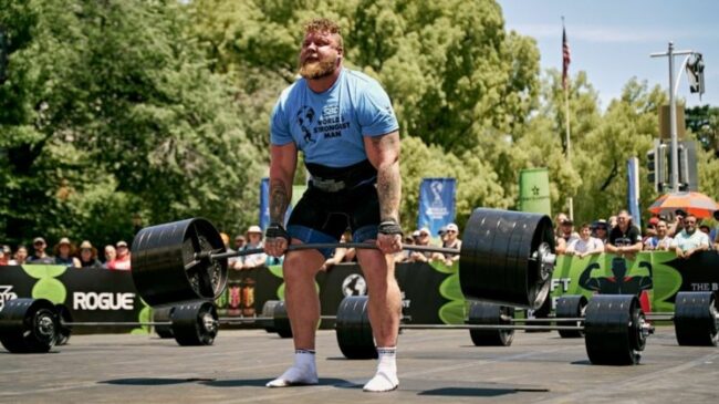 Tom Stoltman, wearing a blue t-shirt and black shirts, deadlifts a barbell loaded with weight plates