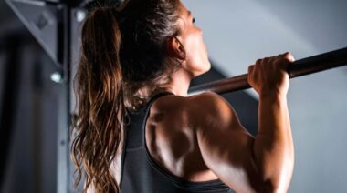 A person with a ponytail wears a sports bra while holding their chin over the bar at the top of their pull-up.